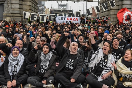 people raise fists and shout, wearing shirts that say ‘not in our name’ and ‘cease fire now’, near signs saying ‘let gaza live’