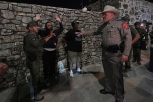 Two men stand against a stone wall, one of them with his hands in the air as a police officer in a stetson addresses him