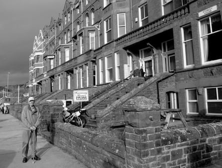 A black and white image of a row of large terraced houses with an elderly man standing on the pavement