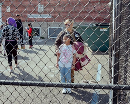Seen through a chain-link fence, a middle-aged Black woman with a frosted blond bob poses standing behind a Black girl of about 10 in T-shirt and jeans, gently holding her arms.