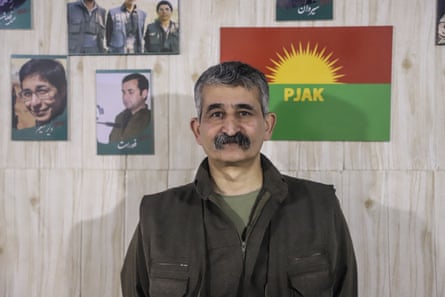 Haftan stands in front of a wall with pictures and a green and red flag with a rising sun on it above the letter PJAK