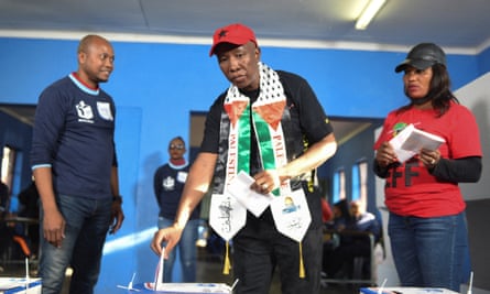 Julius Malema, wearing a Palestinian scarf, puts his vote in a ballot box flanked by two people 