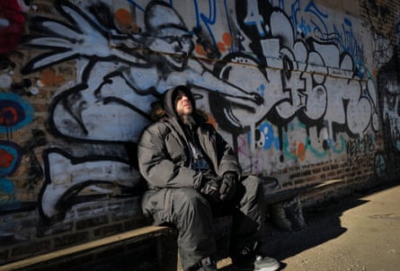 Man sits on a bench in front of a graffiti covered wall in the sunshine.