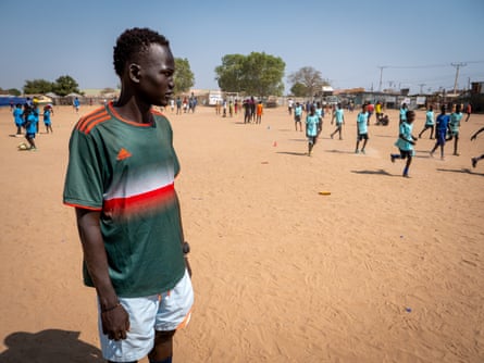 A ypoung man stands on a sand football pitch watching boys play a game