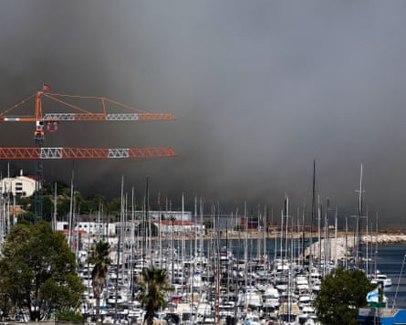 Yachts moored in a harbour with thick black smoke filling the sky behind them.