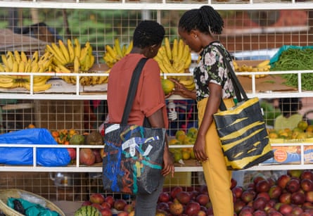 Two people look at fresh produce carrying shopping bags made from plastic waste over their shoulders.