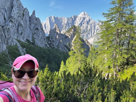 A selfie of Sarah with high craggy peaks, forest and blue sky behind