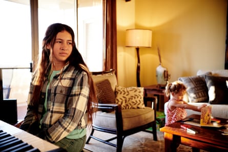A young girl with long brown hair wearing a white checked shirt over a teal T-shirt looks to her left as she sits at a piano keyboard, and a small girl with a curly pigtail in pink plays with something on the low table behind her.