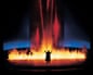 A silhouetted figure stands before a wall of bright orange flames and red light beams on a stage