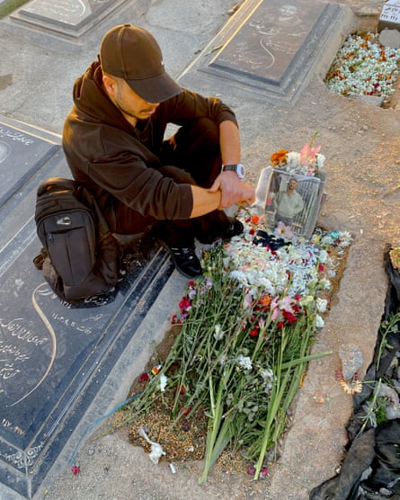 Seen from above, a young man in a baseball cap sits by his friend’s grave.