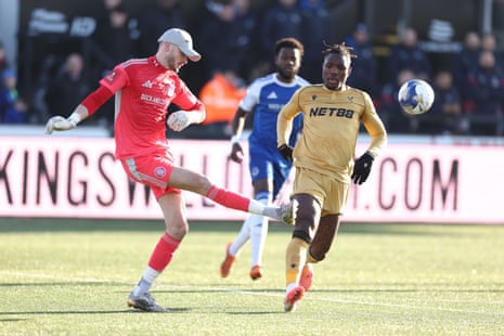 Macclesfield keeer Max Dearnley, sporting a very necessary cap, hoicks the ball clear from the approaching Chrisantus Uche.