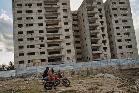 A man and a woman sit on a motorbike on wasteland in front of an unfinished block of flats