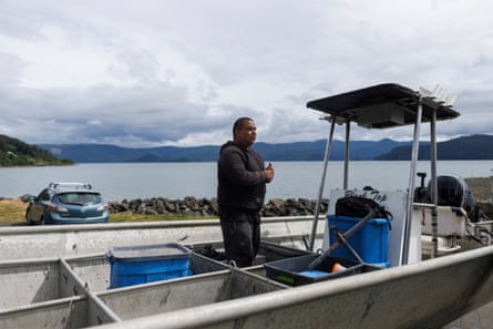 A man stands on a small boat with a bay fringed by mountains in the background