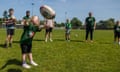 Lucie-Ann, an eight-year-old girl with blond hair, reaches her arms out to catch a rugby ball; she is seen in the foreground on the field with other children and young people of varying ages, and the female coach and an adult player are standing on. They are mostly wearing dark green T-shirts with the Leicester Tigers logo on the front.