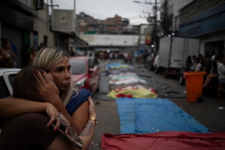 Two women hug close to a line of covered bodies.
