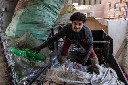 A young man stands in a crate full of plastic drinks bottles, next to large sacks filled with plastic bottles