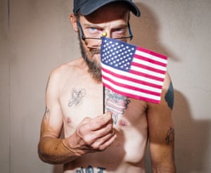 A bare-chested, tattooed man in baseball cap holds a mini American flag