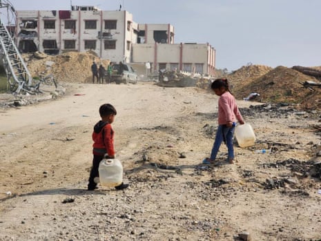 Two young children carry plastic water containers. They are standing on dusty ground with debris and destruction around them.