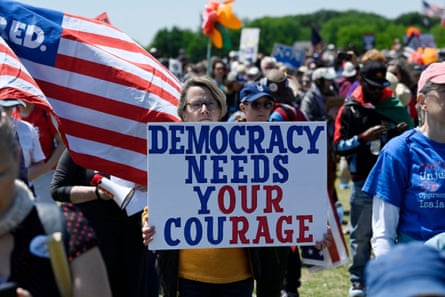 A person holds a sign that reads ‘democracy needs your courage’ among a crowd of protesters and a US flag.