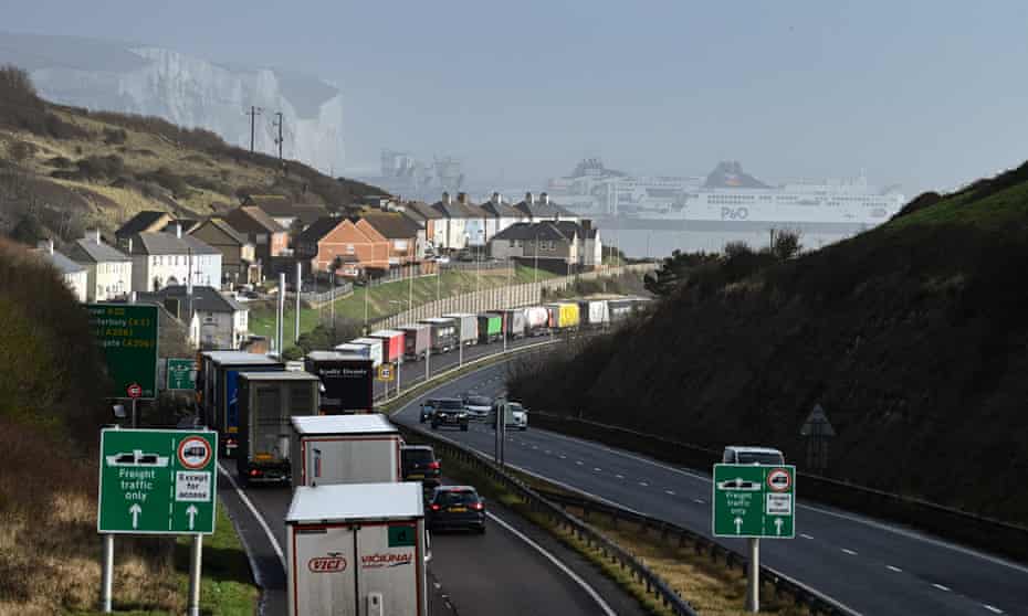A long queue of trucks seen from behind on a road winding down into Dover, wirth the white cliffs and Channel ferries visible in the background