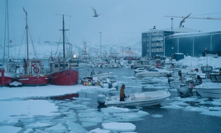 Person in dinghy with two red fishing boats behind and buildings by icy water