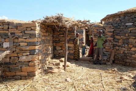 A man and three children stand in the doorway of a stone house, with straw on the floor of the yard