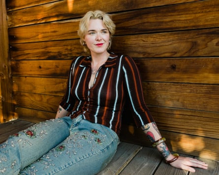 In a striped shirt and jeans, Wedl sits on a porch, leaning back against a wood-panelled wall, looking relaxed