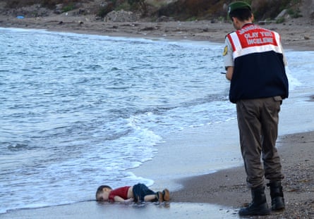 A Turkish police officer stands on a beach near Bodrum, looking down at the body of a child lying face down, dead, at the water’s edge, September 2 2015