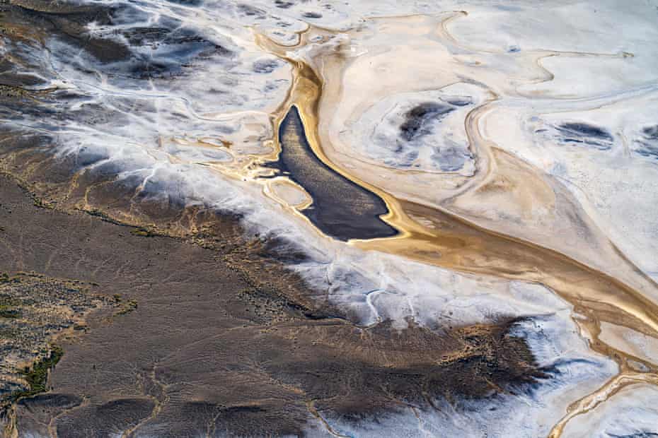 Aerial view after sunrise, showing the sun reflecting off water that has flowed into Lake Eyre South and formed a pond; the water being the result of unusually high local desert rainfall. Visible salt pan edges and streams around edge as well as desert greenery (rare). South Lake Eyre, South Australia, Australia. March 2022