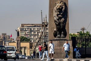 People walk on a bridge under a statue of a lion with cars driving past and a city in the background