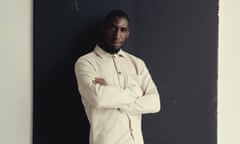 A black man wearing a white shirt folds his arms and looks to the camera, as he stands in front of a black screen