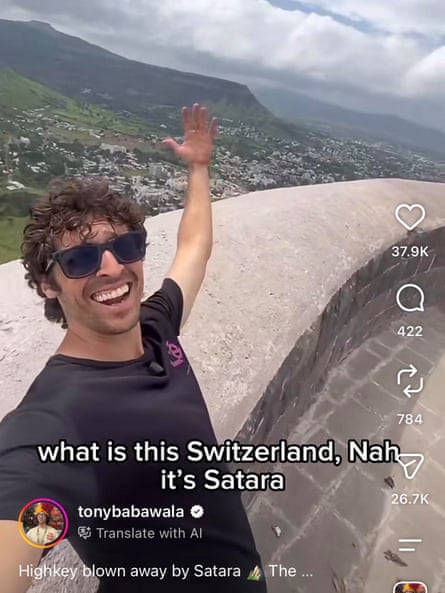 a man on instgram taking a selfie in front of a green city with mountains and clouds in the background