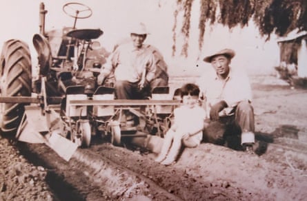 Sepia-tint photo of a Japanese family in a field, posing at the back of a tractor attached to a tiller. A man kneels in a deep rut beside a young child with a bowl cut, who sits on top of the rut, and another man stands against the back of the tiller. All are smiling.