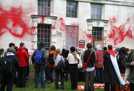 A crowd of young people, seen from behind, gather outside the Ministry of Defence, where red paint has been sprayed on the white stone walls. One of the supporters has a Palestinian flags draped over her shoulders and another is holding a Socialist Worker placard that reads Freedom for Palestine.