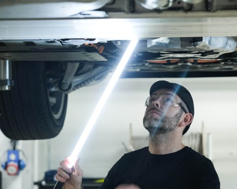 A worker beneath a raised car shines a powerful light on to it