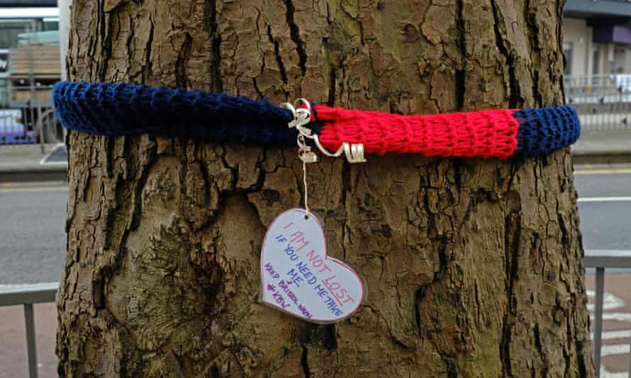 ‘I Am Not Lost’ written on a heart pendant hanging from a blue and red scarf which is tied around a tree.
