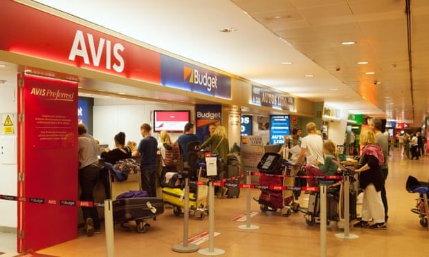 Travellers with luggage queuing at a row of car rental companies' desks in an airport