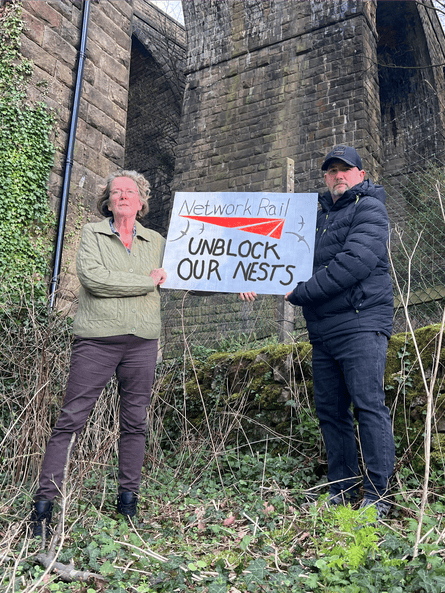 Deb Pitman and Jason Adshead hold a sign saying 'Network Rail, unblock our nests' at the base of a brick viaduct