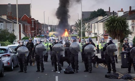 Riot police block protesters in a residential street and near to a burning vehicle.