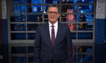 Man wearing a suit, tie, and glasses speaks to the camera on a late-night television set