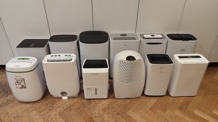 Twelve dehumidifiers, mostly white, black and grey, are lined up on a wooden floor