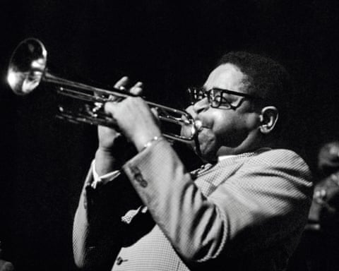Dizzy Gillespie blows a trumpet, cheeks puffed, in a dark stage atmosphere at New York's Apollo theatre