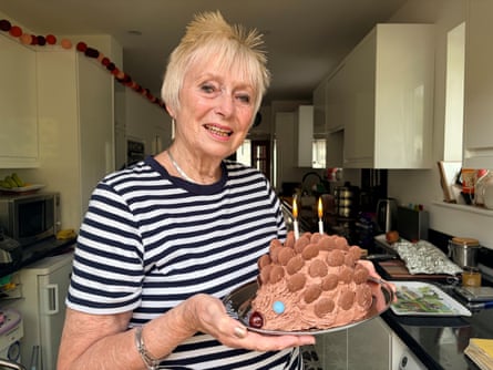 Part of striped T-shirt and a hand with gold nail polish holding a hedgehog birthday cake with chocolate buttons for spines and 2 candles in a modern kitchen