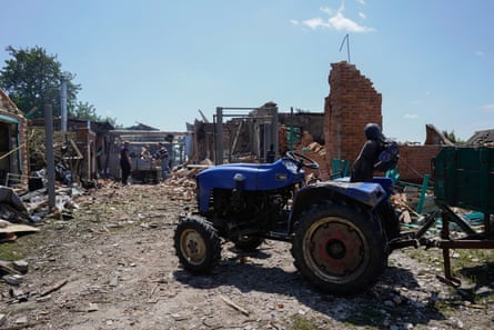 A group of people can be seen in the distance surrounded by rubble, next to the remains of a building. A tractor is in the foreground.