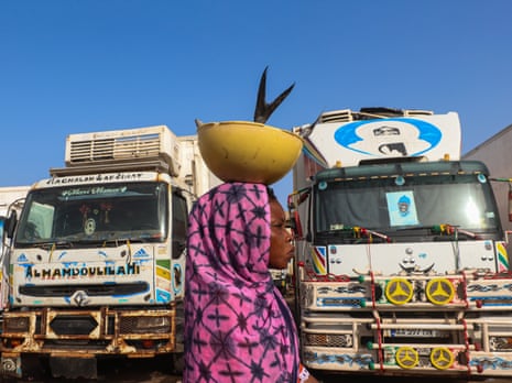 A woman carrying fish in a bowl on her head walks past two trucks