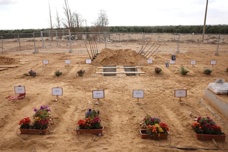 Una hilera de tumbas en la tierra desnuda de un nuevo cementerio, cada una con una simple lápida y un ramo de flores rojas al pie de la tumba.