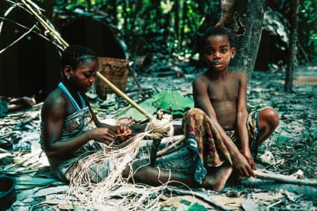 Two children sit on the ground in a rainforest clearing weaving with grass-like fibres.