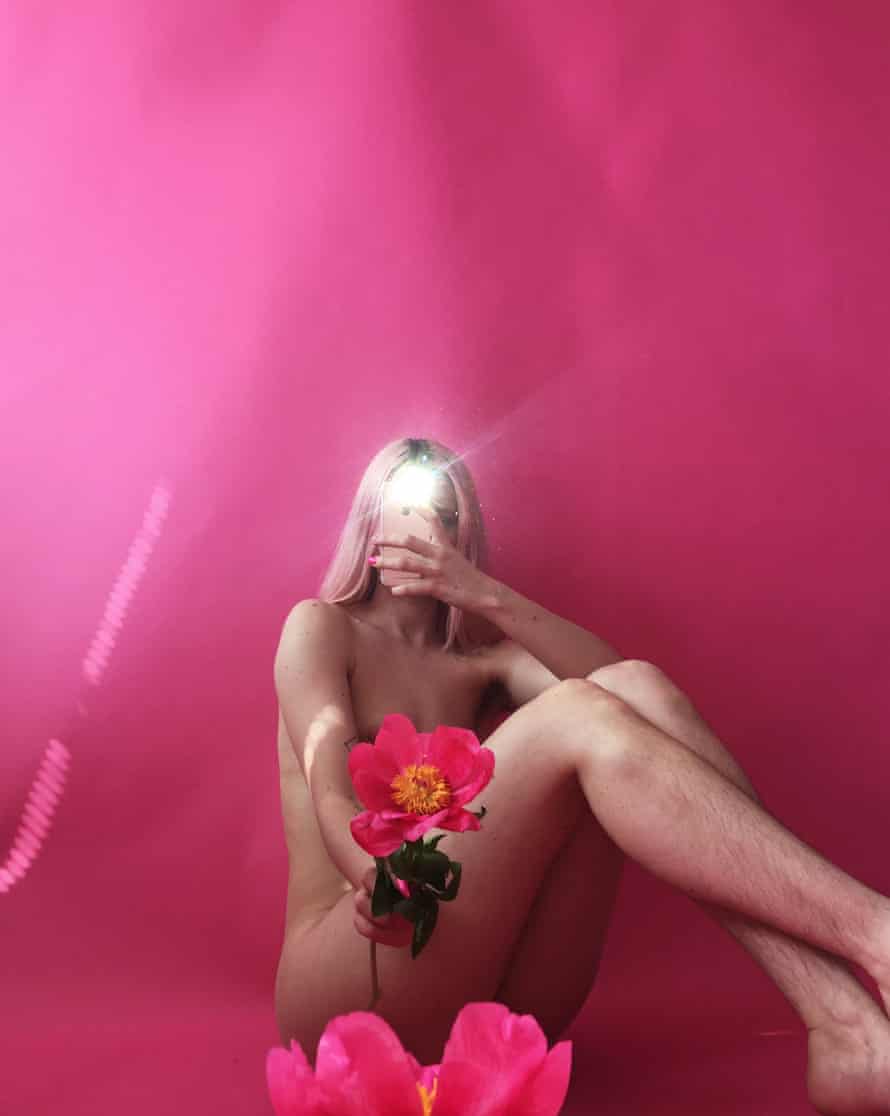 A slender, nude young woman with long blonde hair sits  on the ground in front of a bright pink background, her legs bent at the knee and crossed at the ankle to conceal her genitalia. In one hand she holds out a pink flower toward the camera, in a way that conceals her breasts. In the other hand she holds a smartphone up in a way that conceals her face behind the flash light of her phone’s camera.