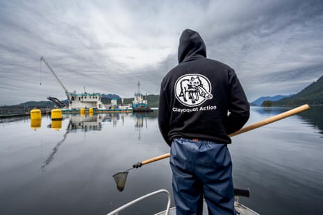 A man in a blue hoodie stands on the pro of a small boat approaching a fish farm on a lake