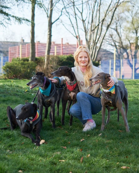 She is crouched down, with her arms around three grey-black greyhounds, a fourth lying on the floor close by